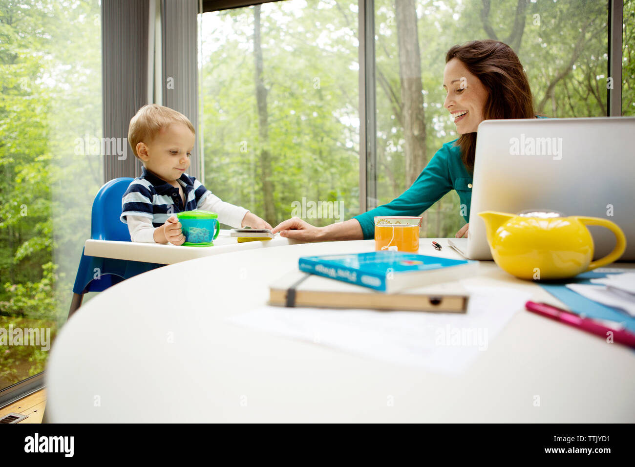 Smiling mother teaching son while using laptop at table against window Stock Photo