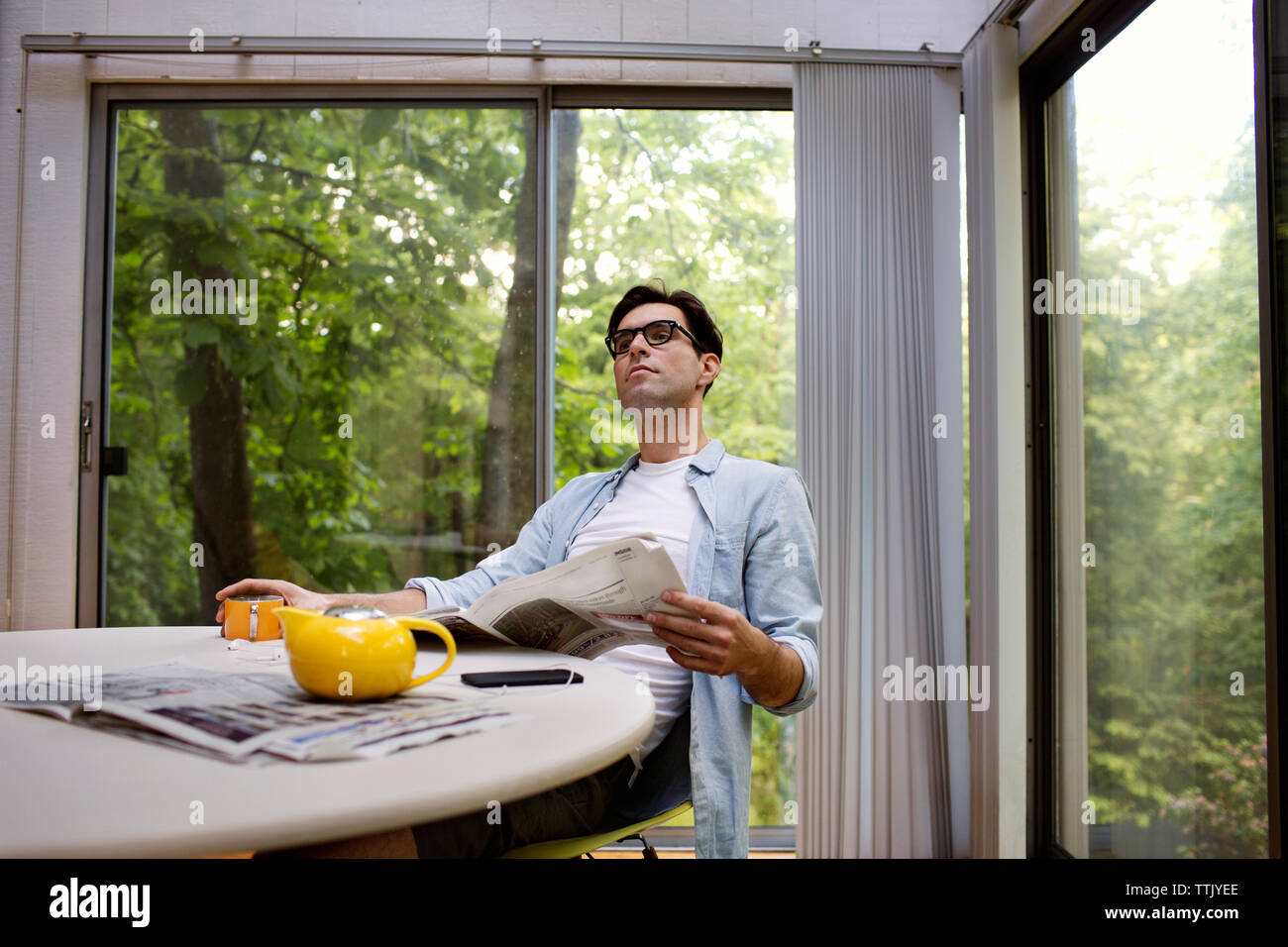 Thoughtful man holding newspaper while having tea at table against window Stock Photo
