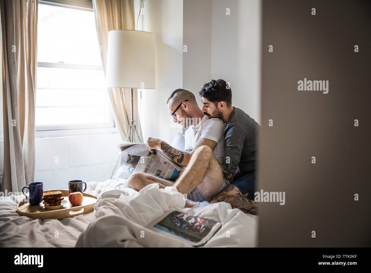 Gay men reading newspaper while sitting on bed at home Stock Photo
