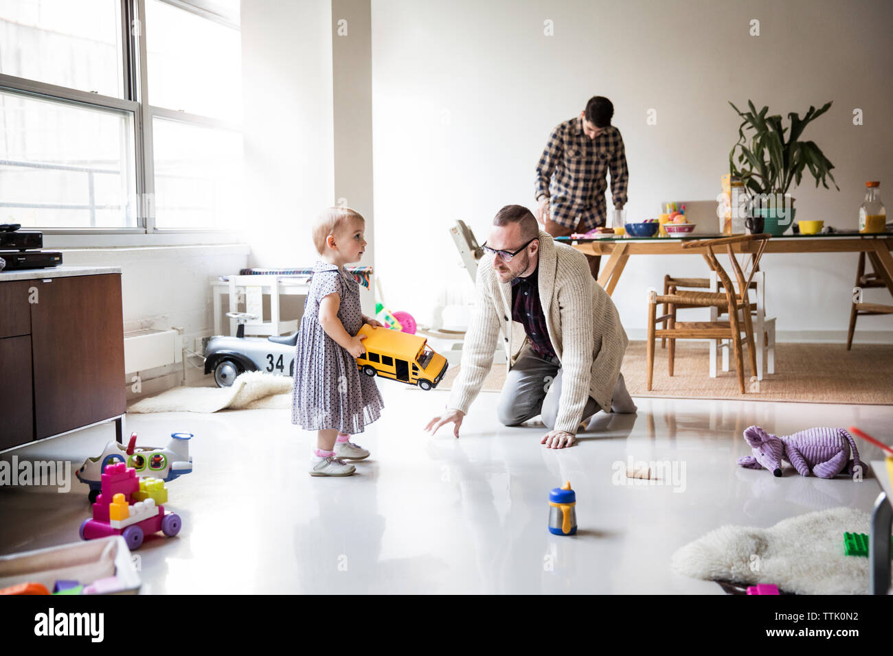 Father playing with daughter with partner working at table in brightly lit living room Stock Photo