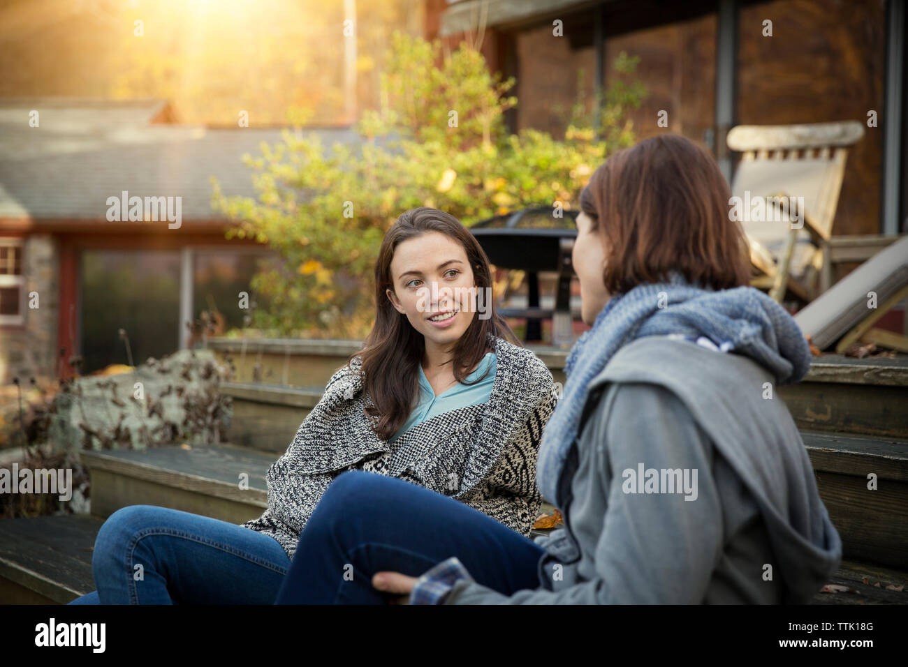 Smiling daughter and mother talking while sitting on steps outside log cabin in forest Stock Photo