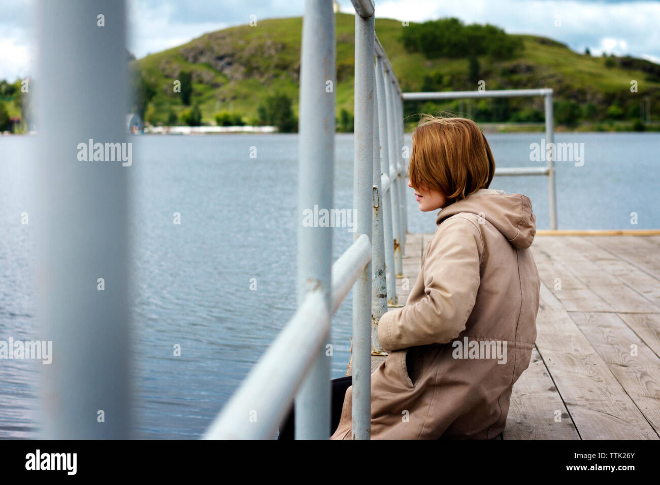 Woman wearing long coat sitting at jetty by railing over lake Stock Photo