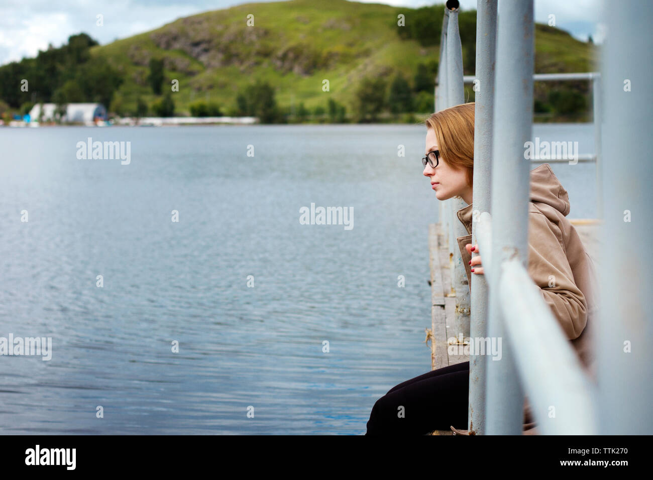 Thoughtful woman sitting at jetty by railing over lake Stock Photo