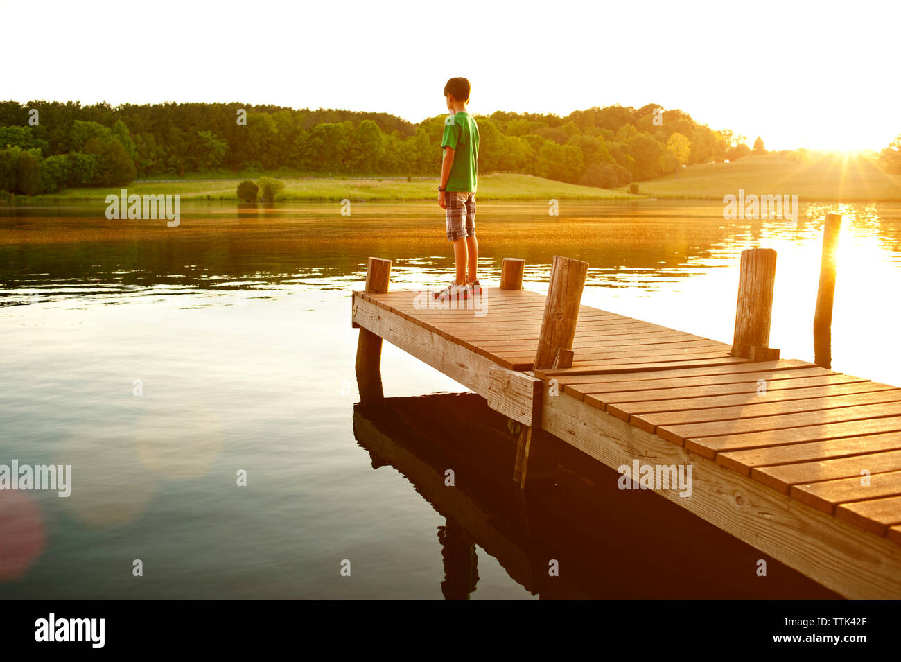Boy standing on jetty over lake during sunny day Stock Photo