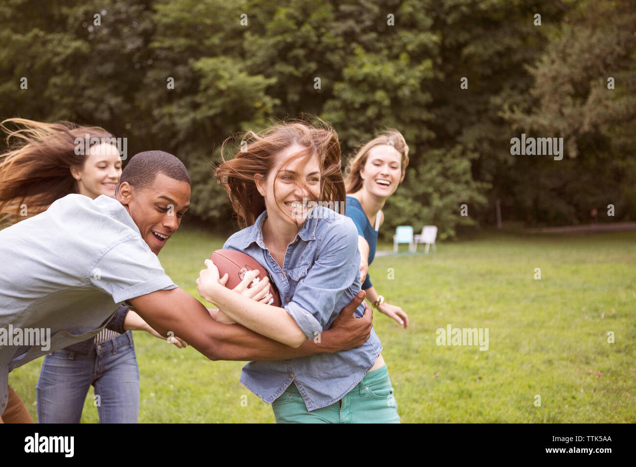 Man pulling woman holding football ball while friends running in background on field Stock Photo
