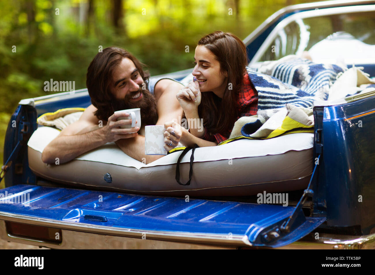 Cheerful couple holding coffee mugs while lying in pick-up truck at forest Stock Photo