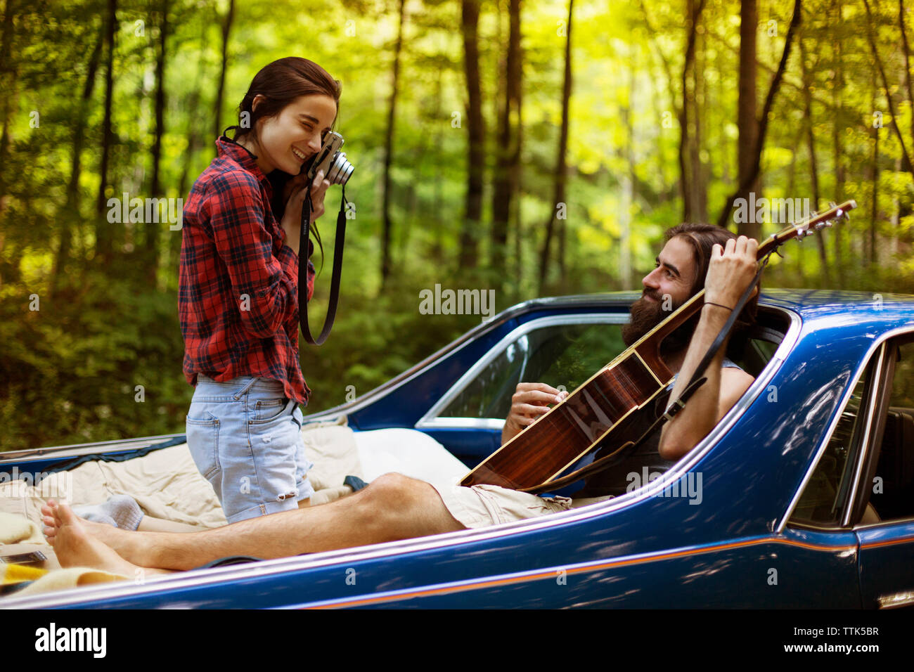 Woman photographing boyfriend holding guitar in pick-up truck at forest Stock Photo