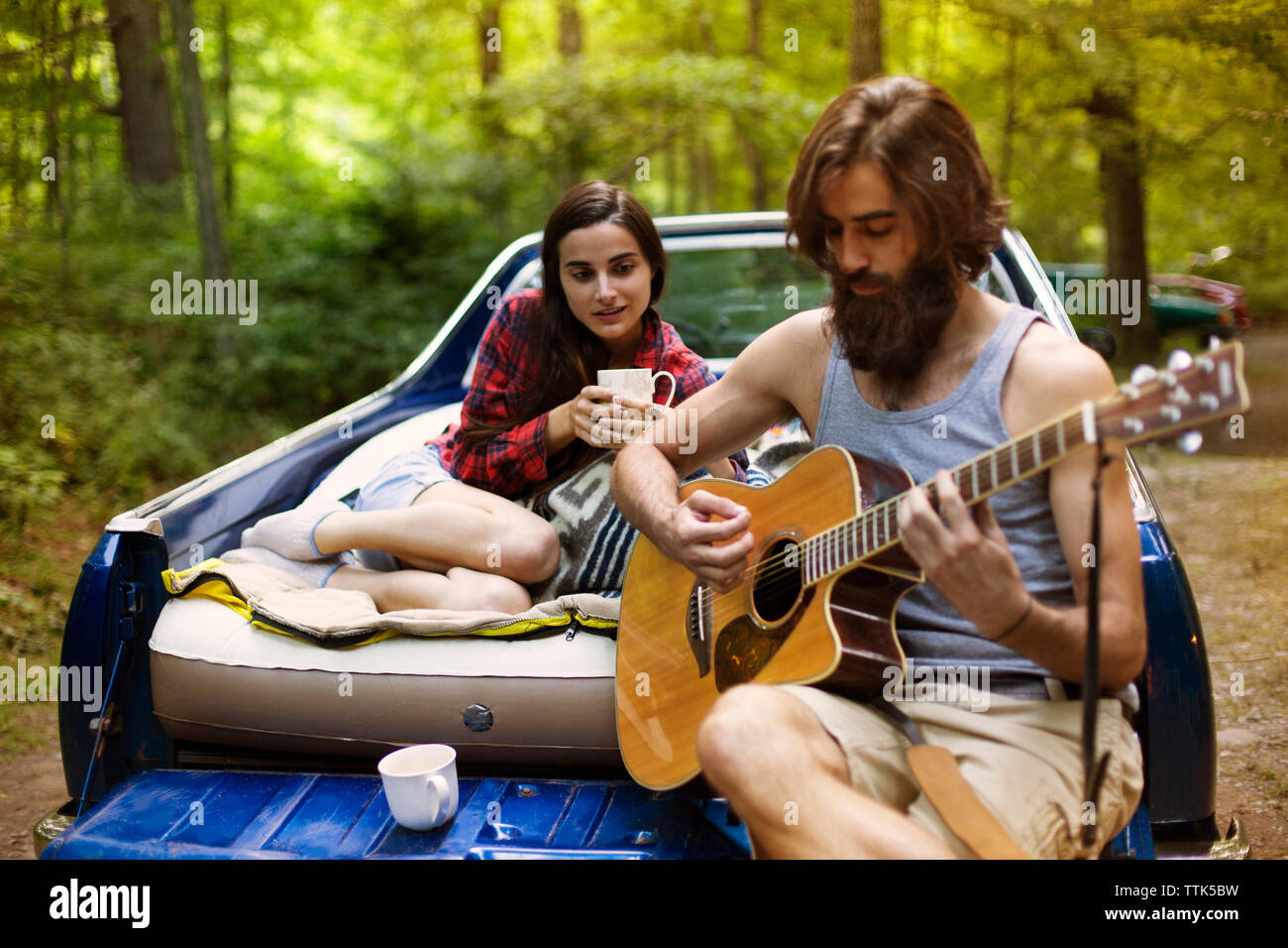 Man playing guitar for girlfriend while sitting in pick-up truck at forest Stock Photo