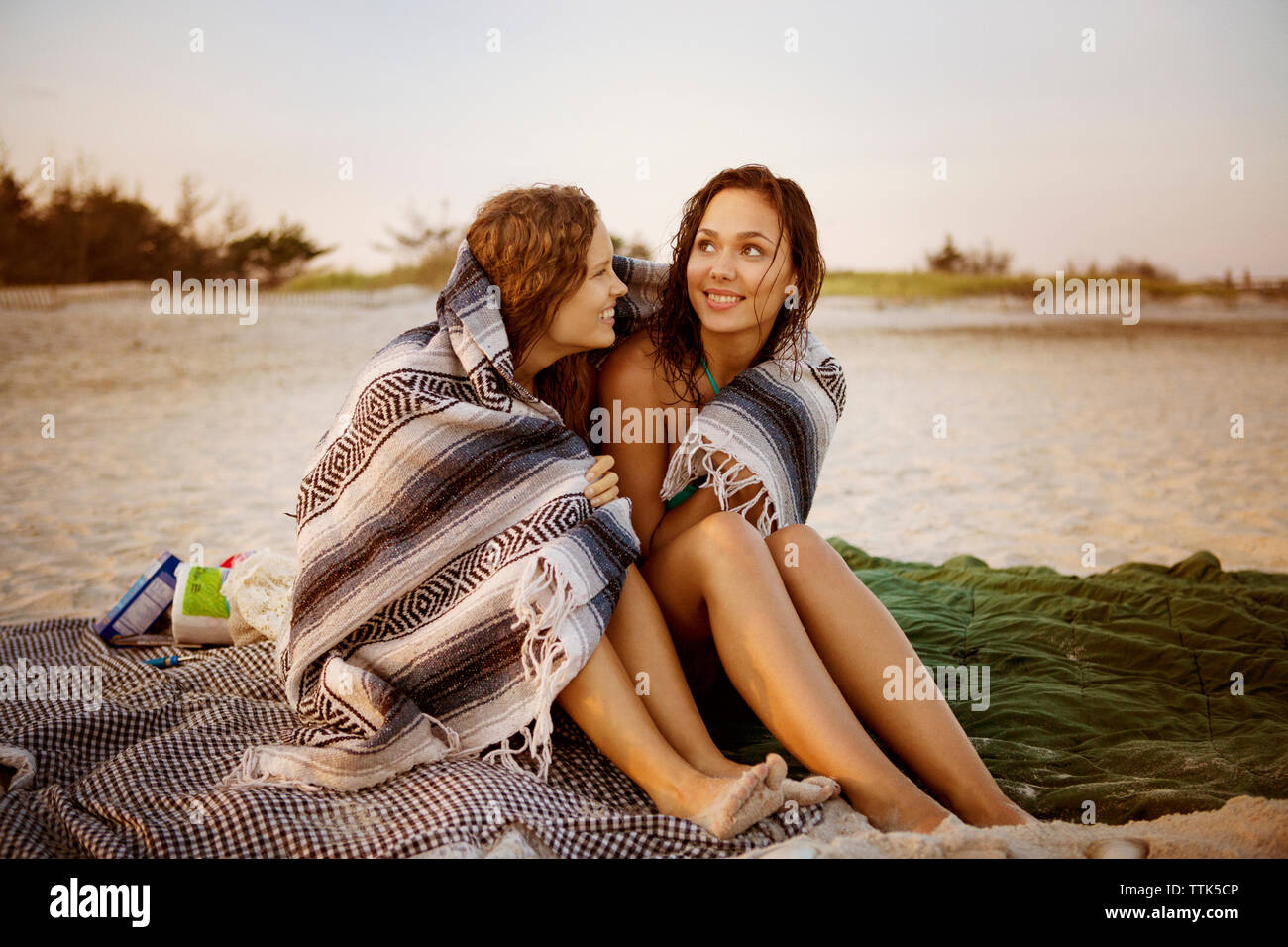 Smiling women wrapped in blanket sitting at beach Stock Photo