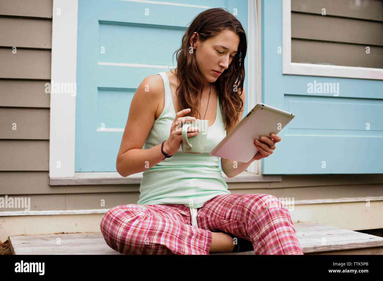 Young woman using tablet computer outside house while holding coffee mug Stock Photo