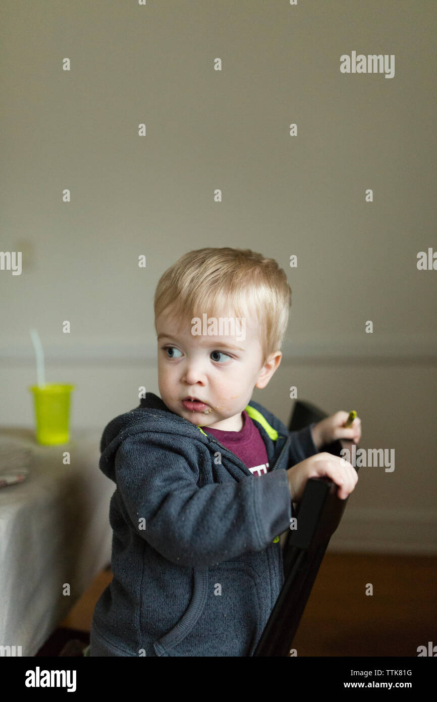 Toddler boy kneeling on chair looks behind him while holding a crayon Stock Photo