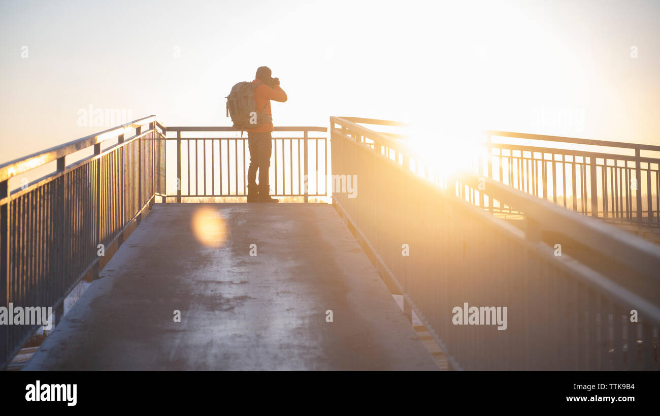 Man taking photos of the sun at sunset on footbridge with backpack Stock Photo
