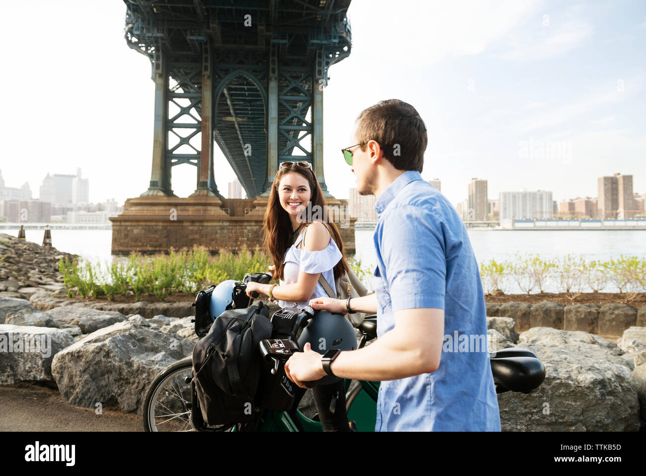Happy couple walking with bicycle by Manhattan bridge against sky Stock Photo