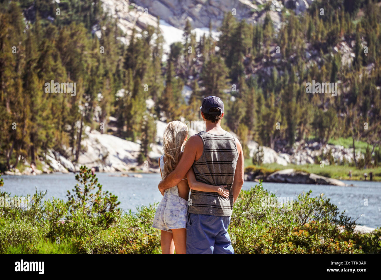 Rear view of couple with arms around looking at lake against mountain while standing in forest during sunny day Stock Photo