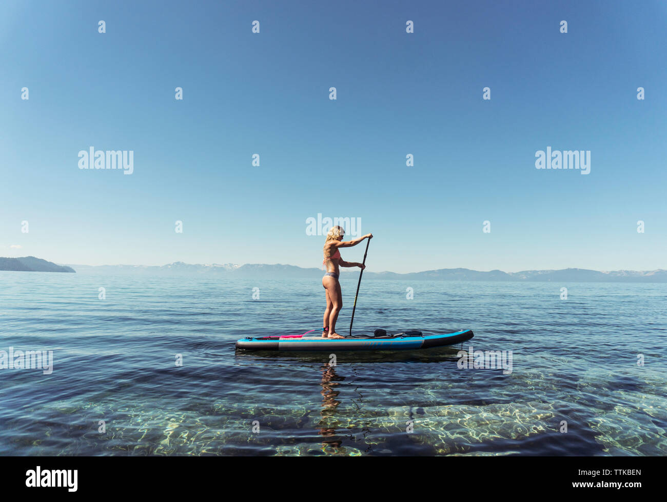 Side view of woman in bikini paddleboarding on lake against clear blue sky during sunny day Stock Photo