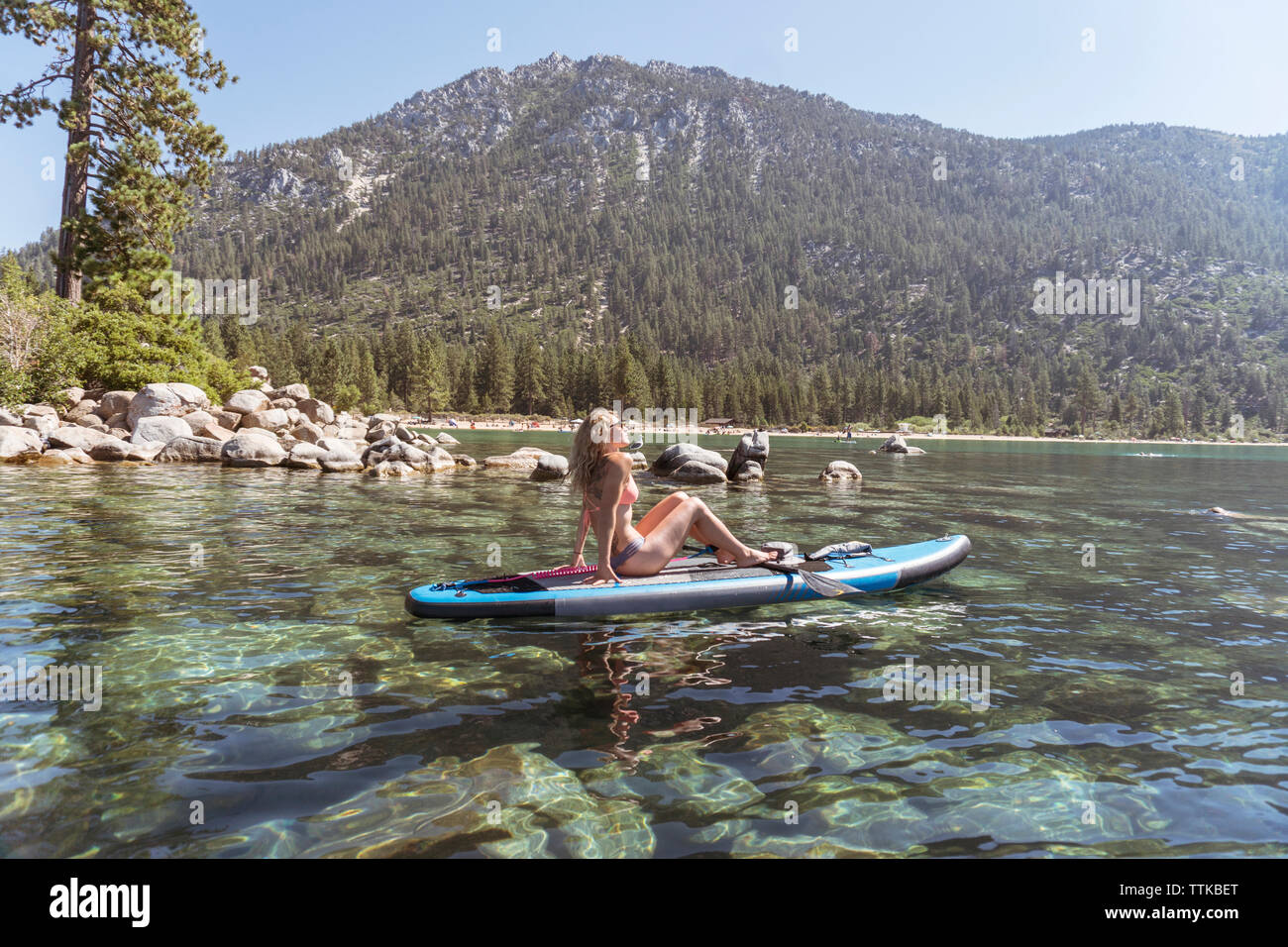Side view of woman in bikini paddleboarding on lake against mountain during sunny day Stock Photo