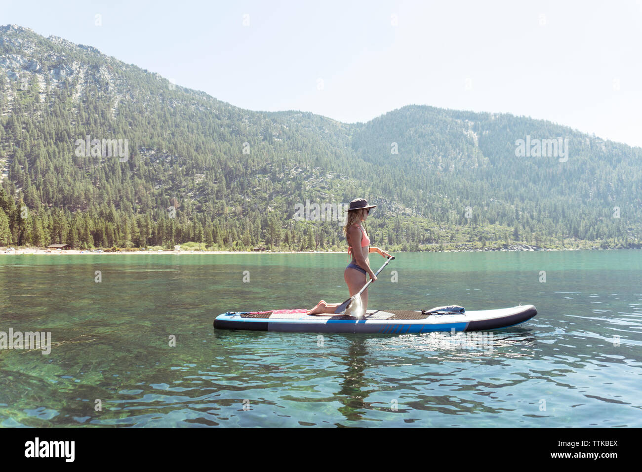 Side view of woman in bikini paddleboarding on lake against clear sky during sunny day Stock Photo