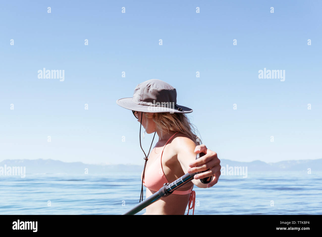 Side view of woman wearing bikini paddleboarding on lake against clear blue sky during sunny day Stock Photo