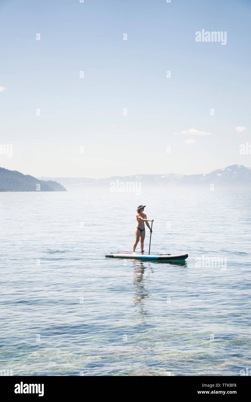 Full length of woman in bikini paddleboarding on lake against blue sky during sunny day Stock Photo
