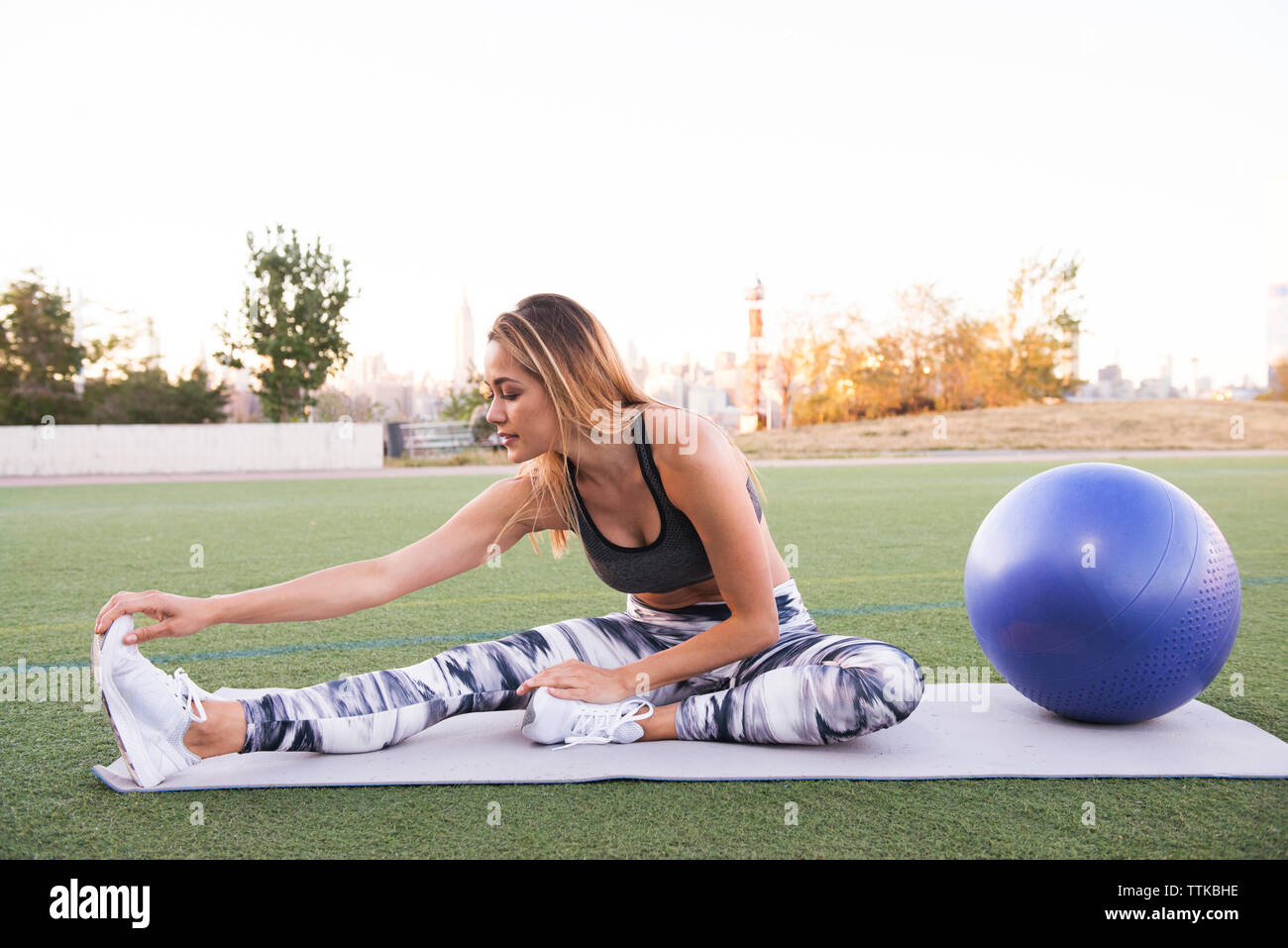 Full length of young woman stretching while sitting on exercise mat by fitness ball against clear sky at park Stock Photo