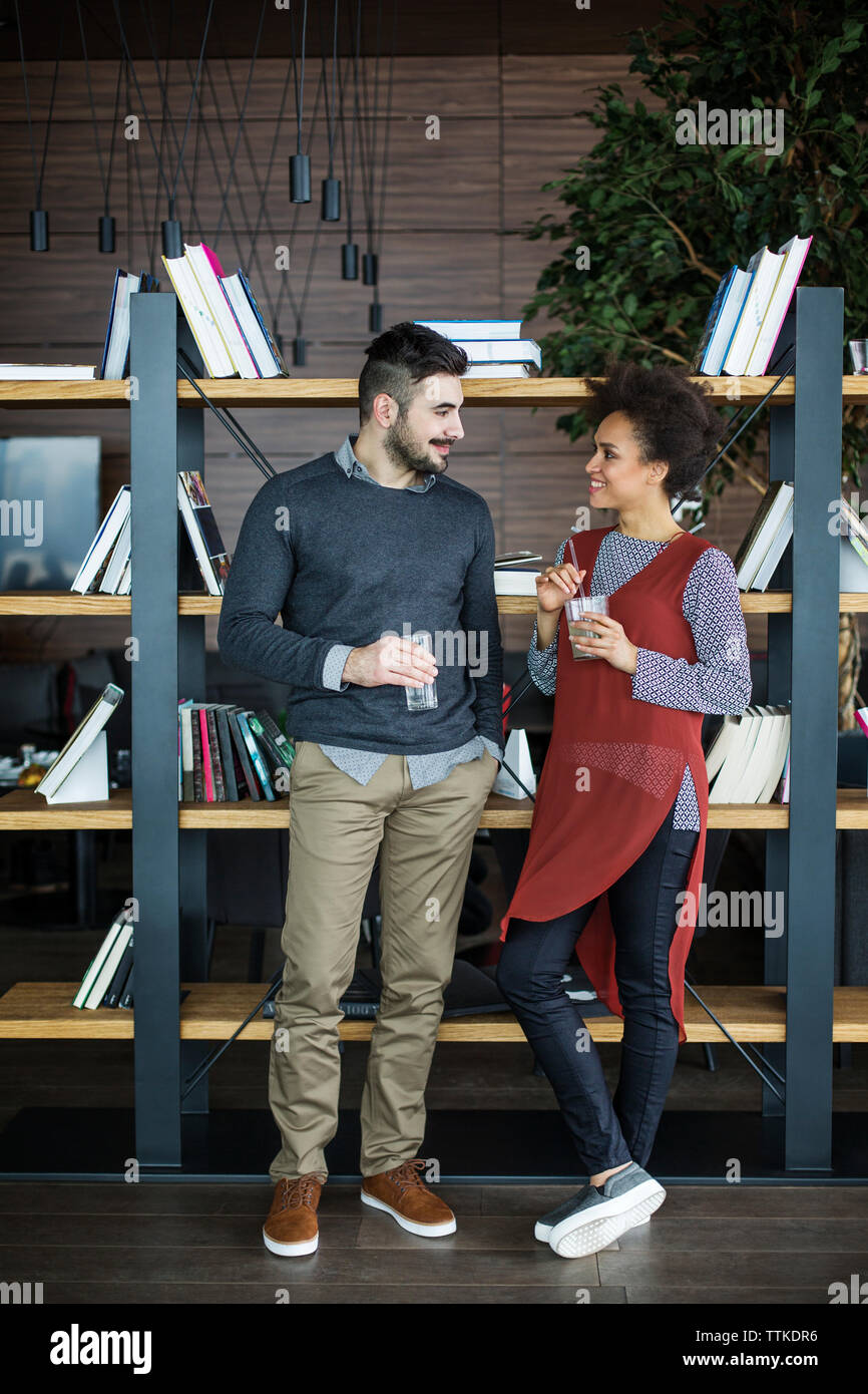 Happy business couple holding drinks while standing against book shelf in restaurant Stock Photo