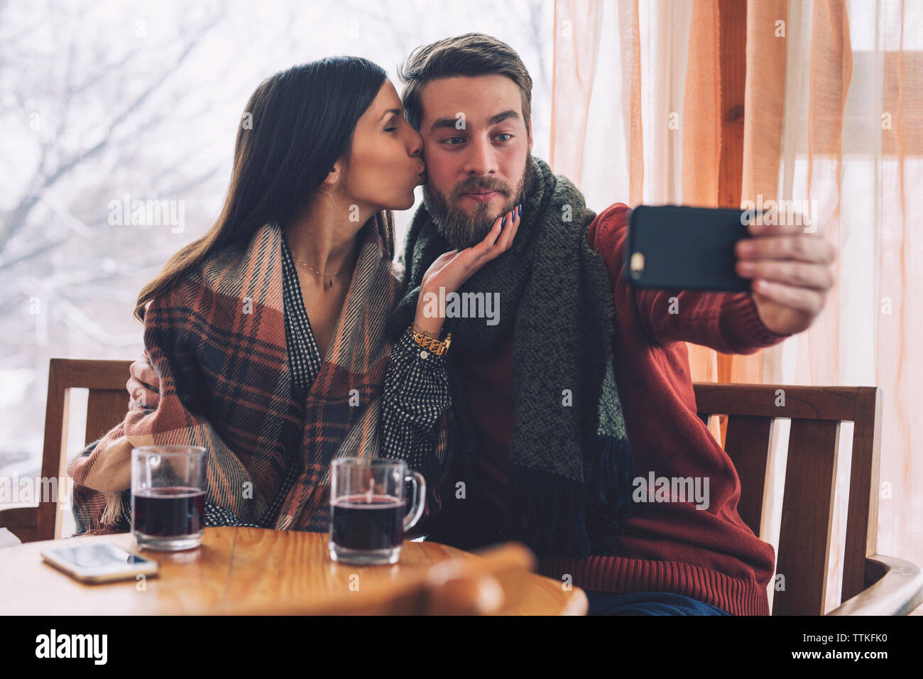 Woman kissing while boyfriend taking selfie at table in cafe Stock Photo