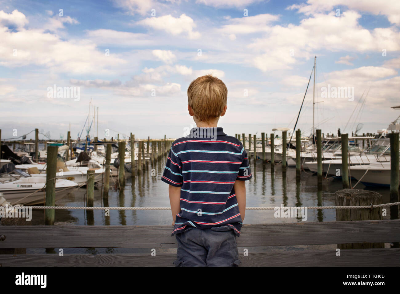 Rear view of boy standing on bridge by harbor against cloudy sky Stock Photo