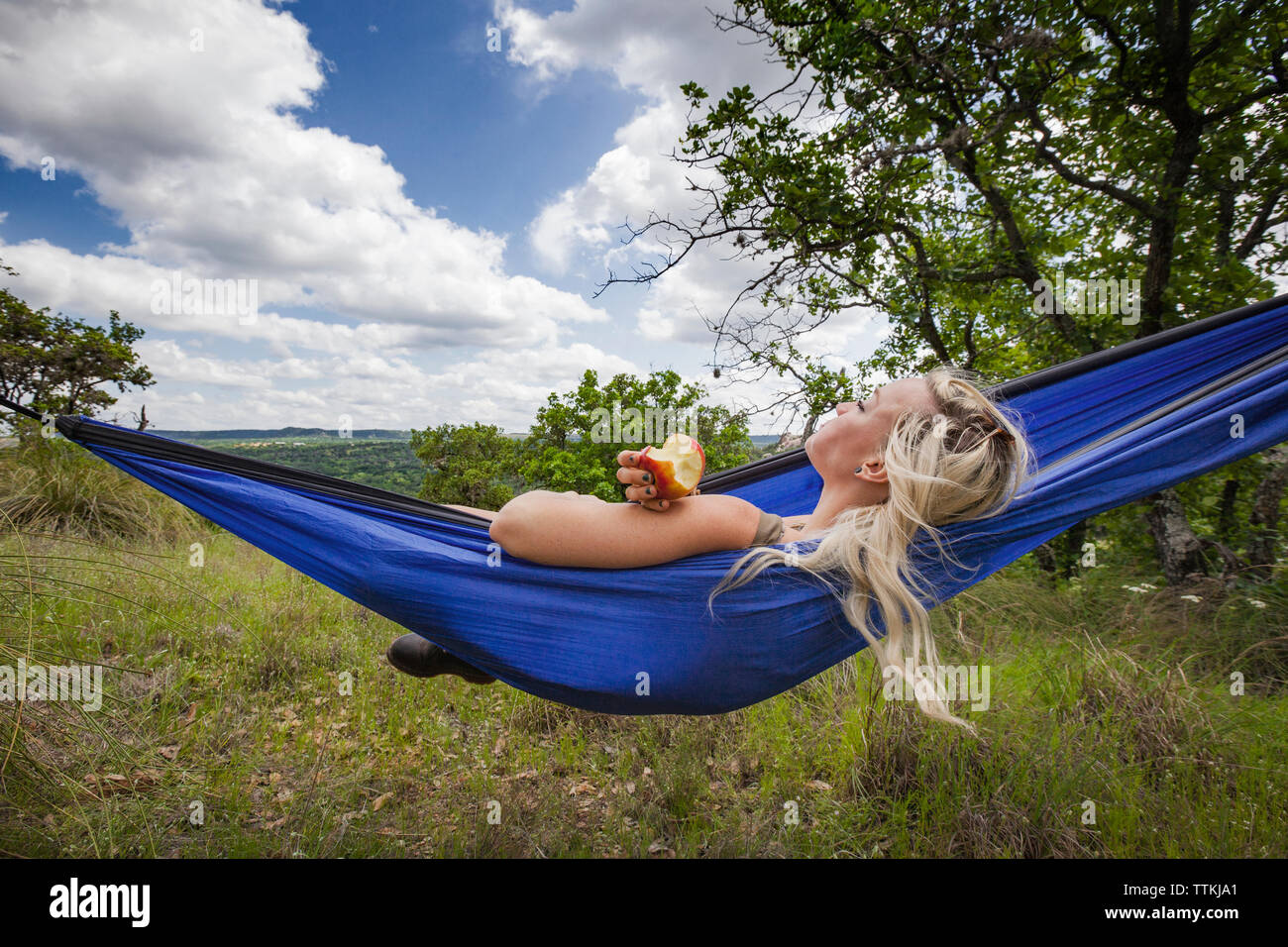 Woman holding apple while lying on hammock at field during vacation Stock Photo