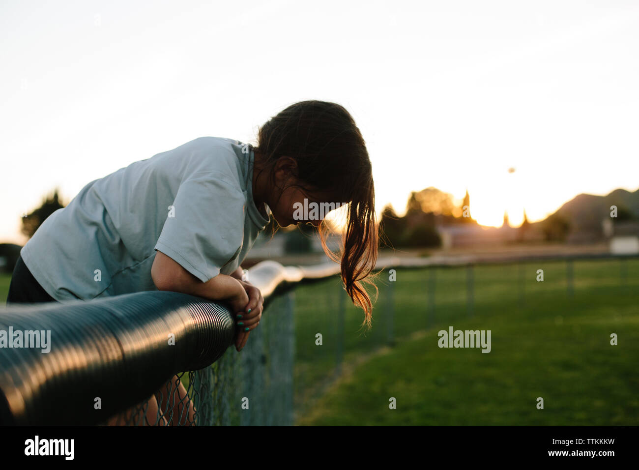 Side view of girl bending over fence at playground during sunset Stock Photo