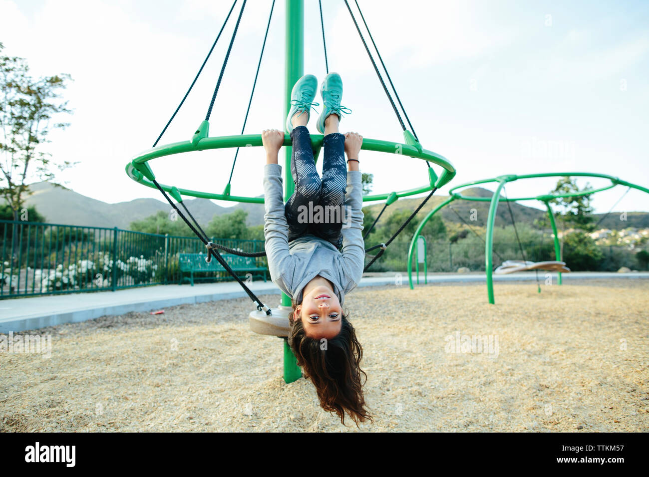Portrait of girl hanging upside down on outdoor play equipment against sky at playground Stock Photo