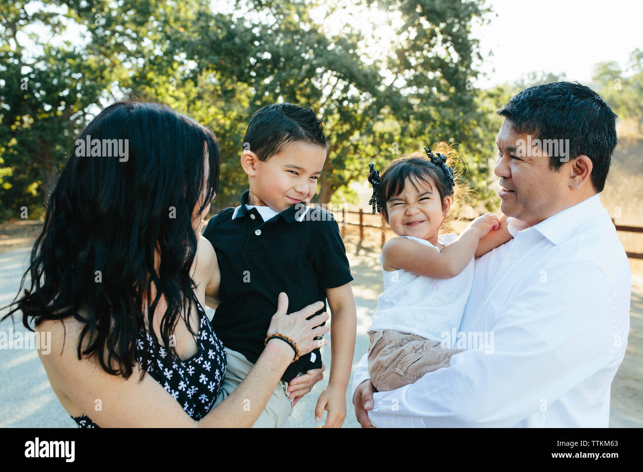 Happy parents carrying cute children making faces while standing on road against trees in park Stock Photo