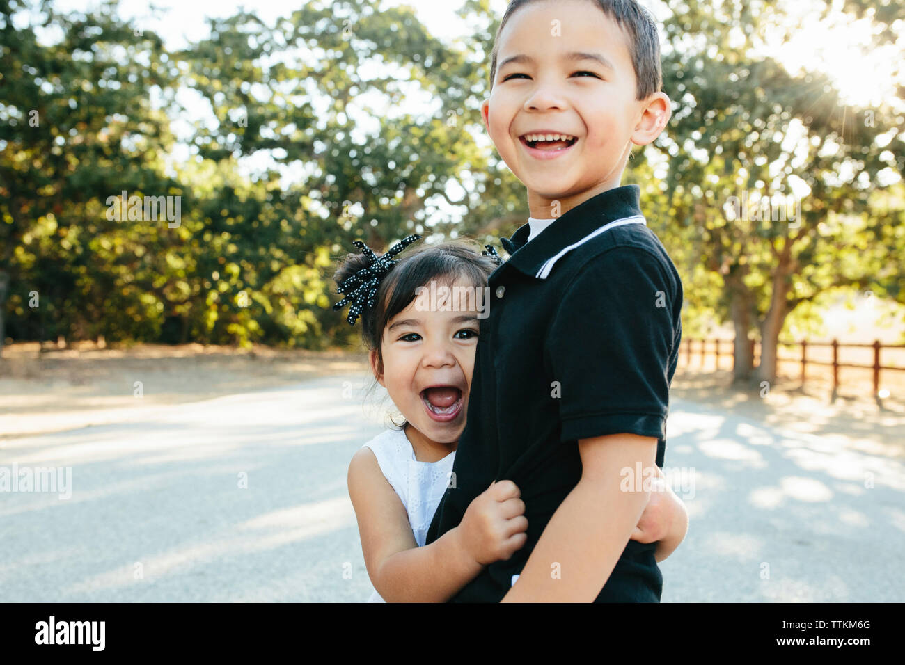 Portrait of happy sister with mouth open embracing brother while standing on road against trees in park Stock Photo