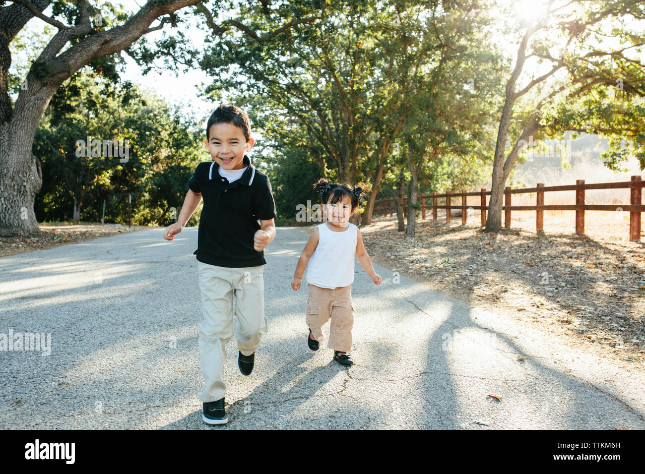 Portrait of happy cute siblings running on road against trees in park Stock Photo
