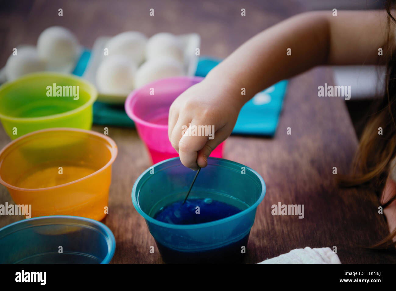 Cropped hand of girl using stirrer to mix dye in water for easter Egg coloring on table Stock Photo