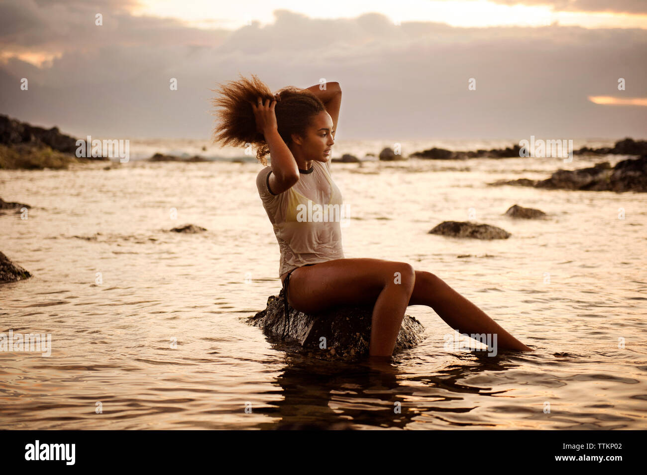 Teenage girl sitting on rock with hand in hair amidst sea Stock Photo