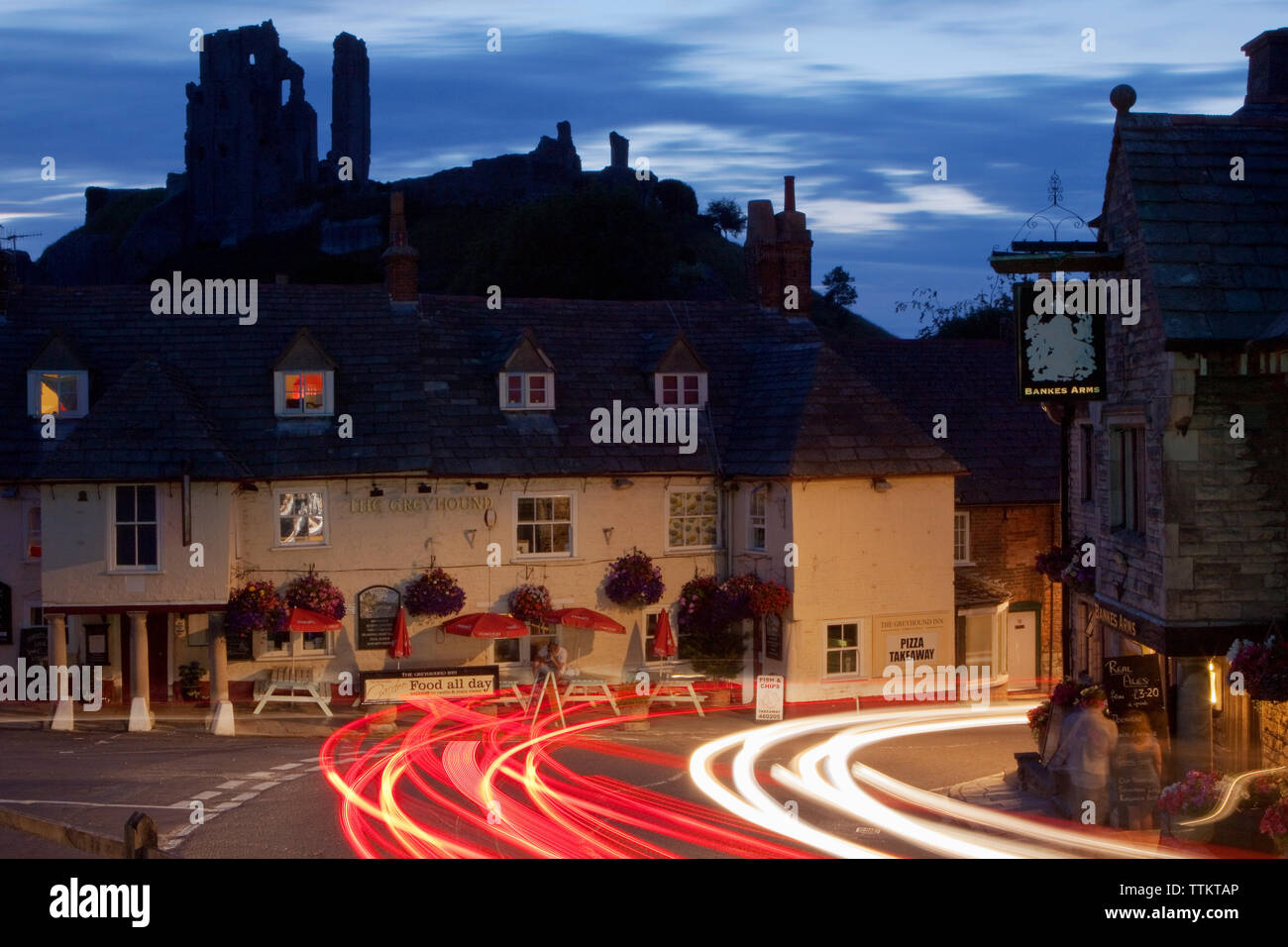 Traffic light trails in a long exposure photograph taken as cars pass The Bankes Arms and The Greyhound pub in front of the ruins of Corfe Castle Stock Photo