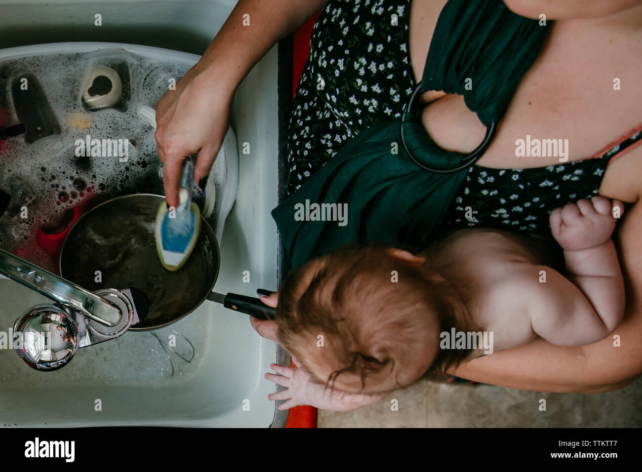Mother carrying newborn child does dishes at home in kitchen Stock Photo