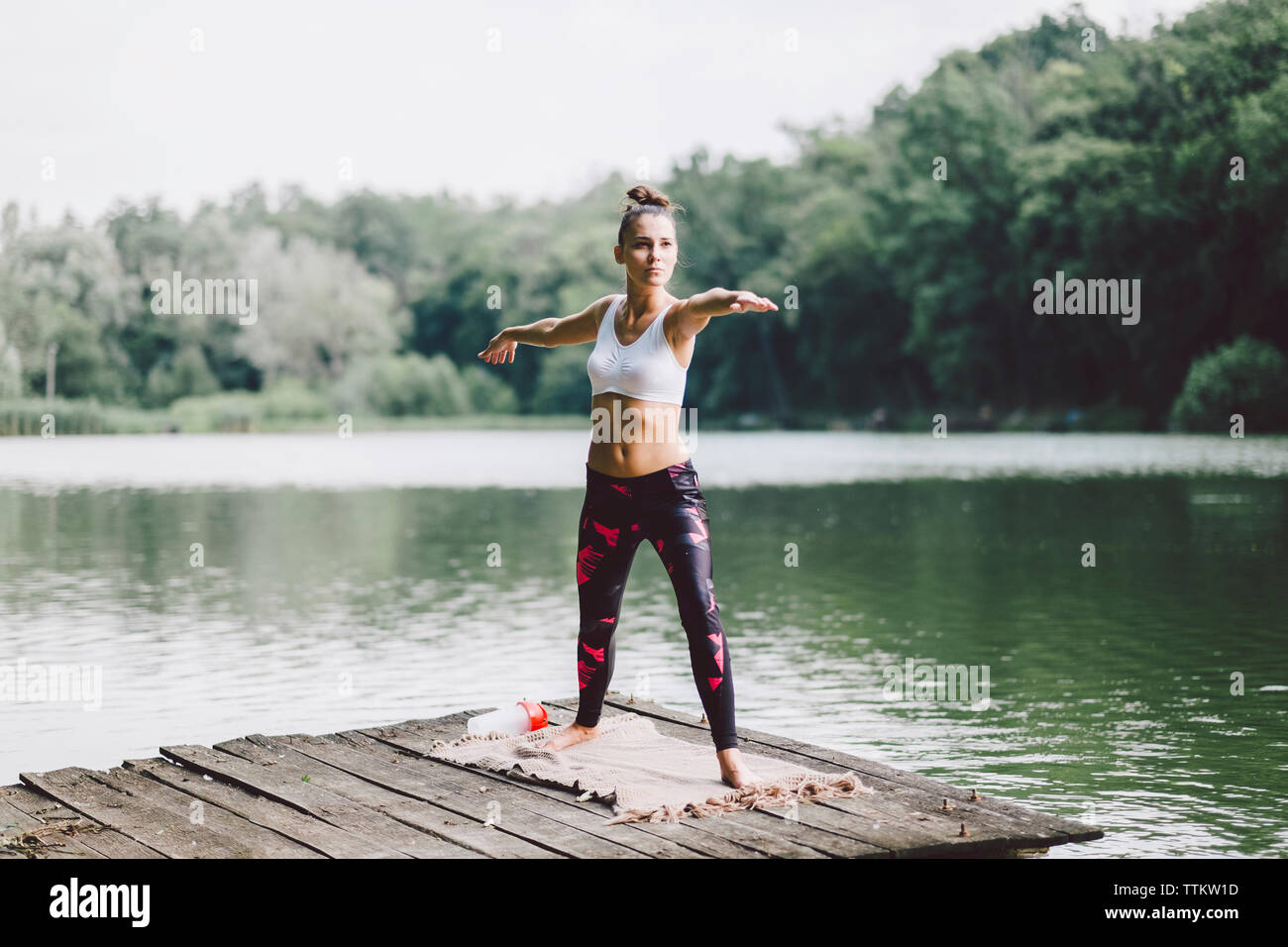 Woman practicing warrior 2 pose while standing on pier over lake in forest Stock Photo