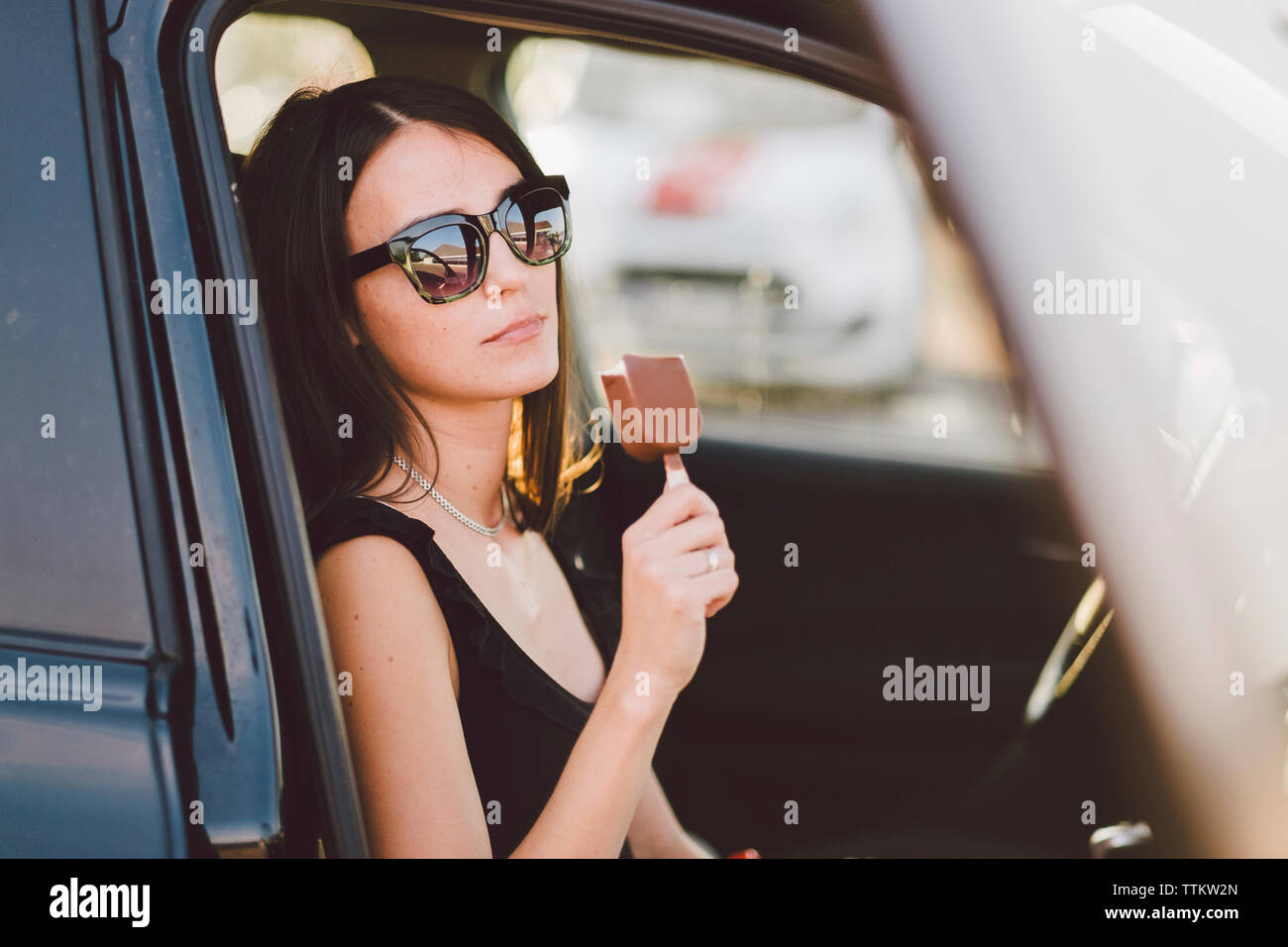 Young woman wearing sunglasses holding frozen sweet food while sitting in car seen through window Stock Photo
