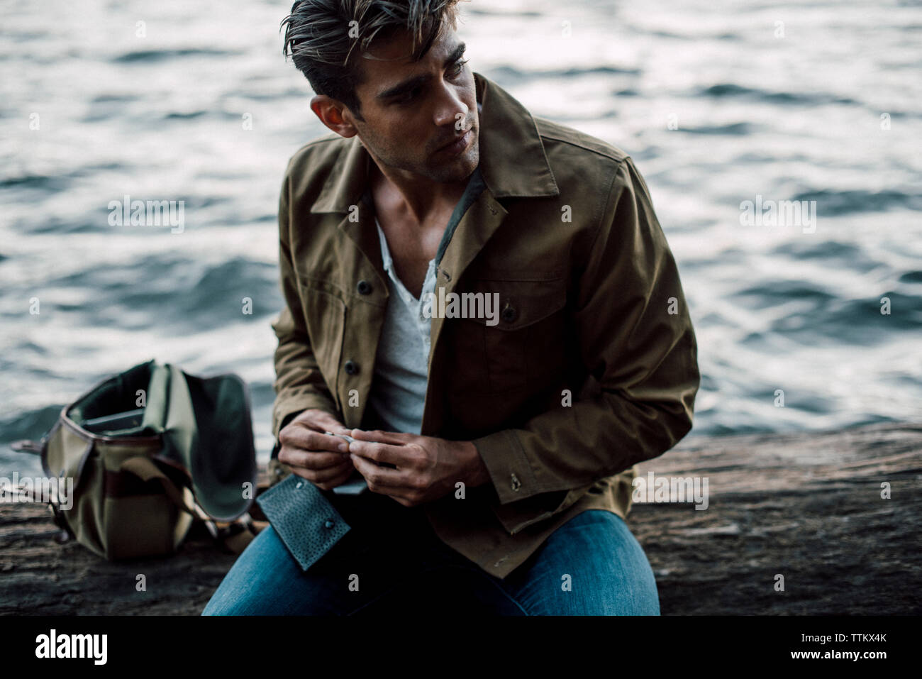 Man holding cigarette looking away while sitting by river on log Stock Photo