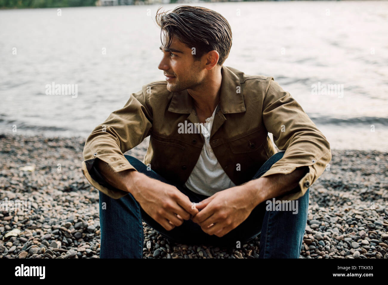 Thoughtful man looking away while sitting at riverbank during sunset Stock Photo