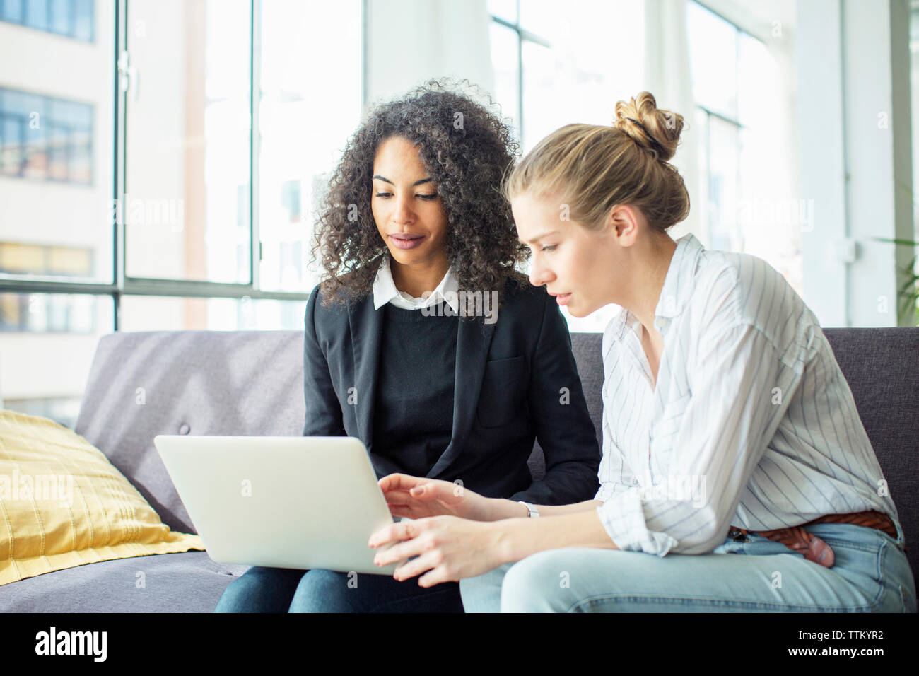 Female colleagues discussing while using laptop computer on sofa in office Stock Photo
