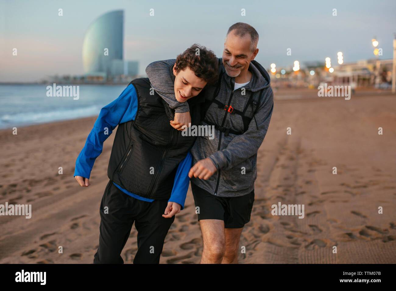 Playful father with arm around son's shoulder walking against hotel Vela at beach Stock Photo