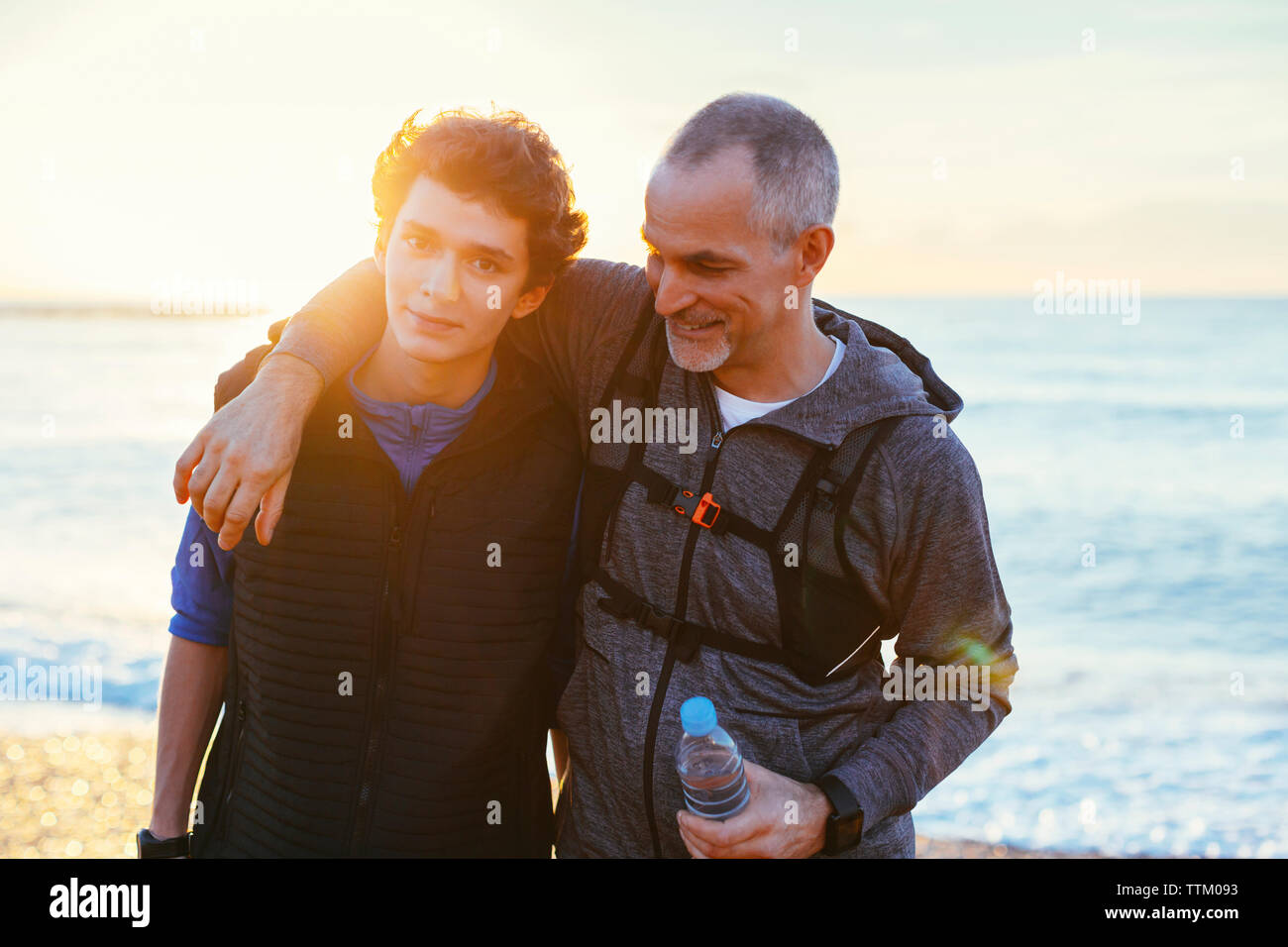 Portrait of son with father's arm around shoulder against sea during sunset Stock Photo