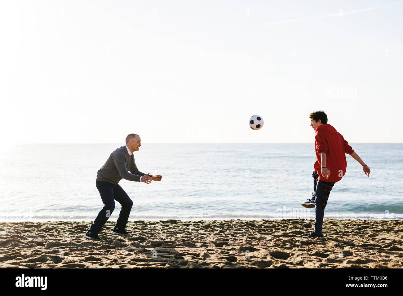Playful son kicking soccer ball while father defending at beach against clear sky Stock Photo