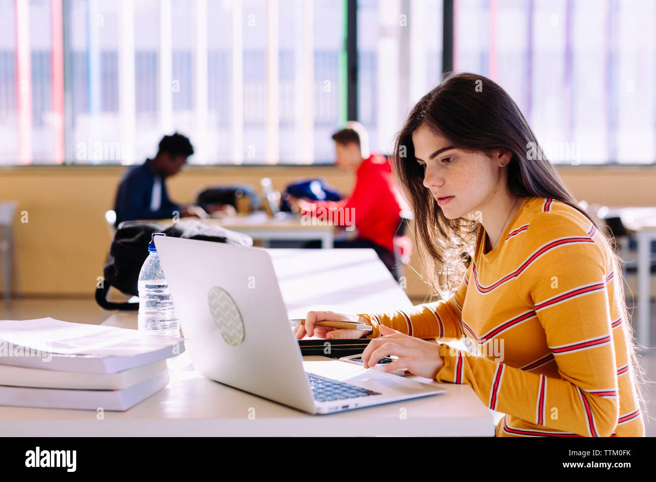 Side view of woman using laptop while male friends studying in background at library Stock Photo