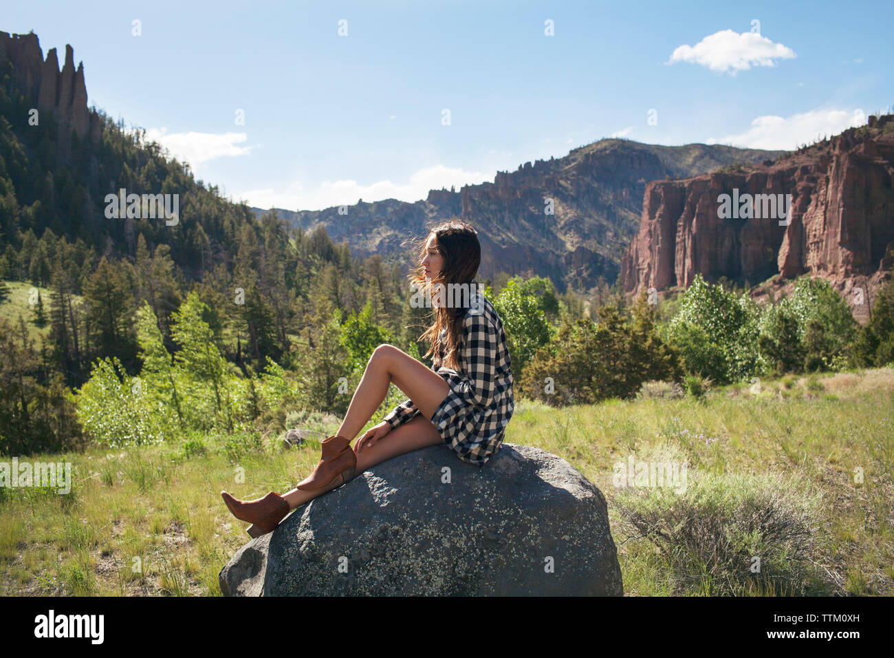Side view of woman sitting on rock at grassy field against mountains Stock Photo