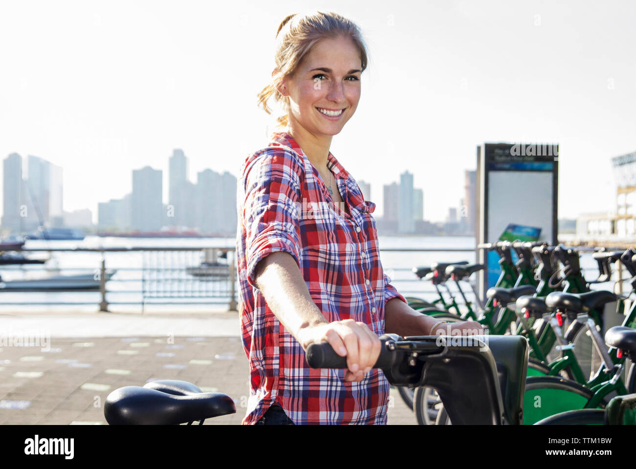 Portrait of confident smiling woman renting bicycle by lake in city Stock Photo
