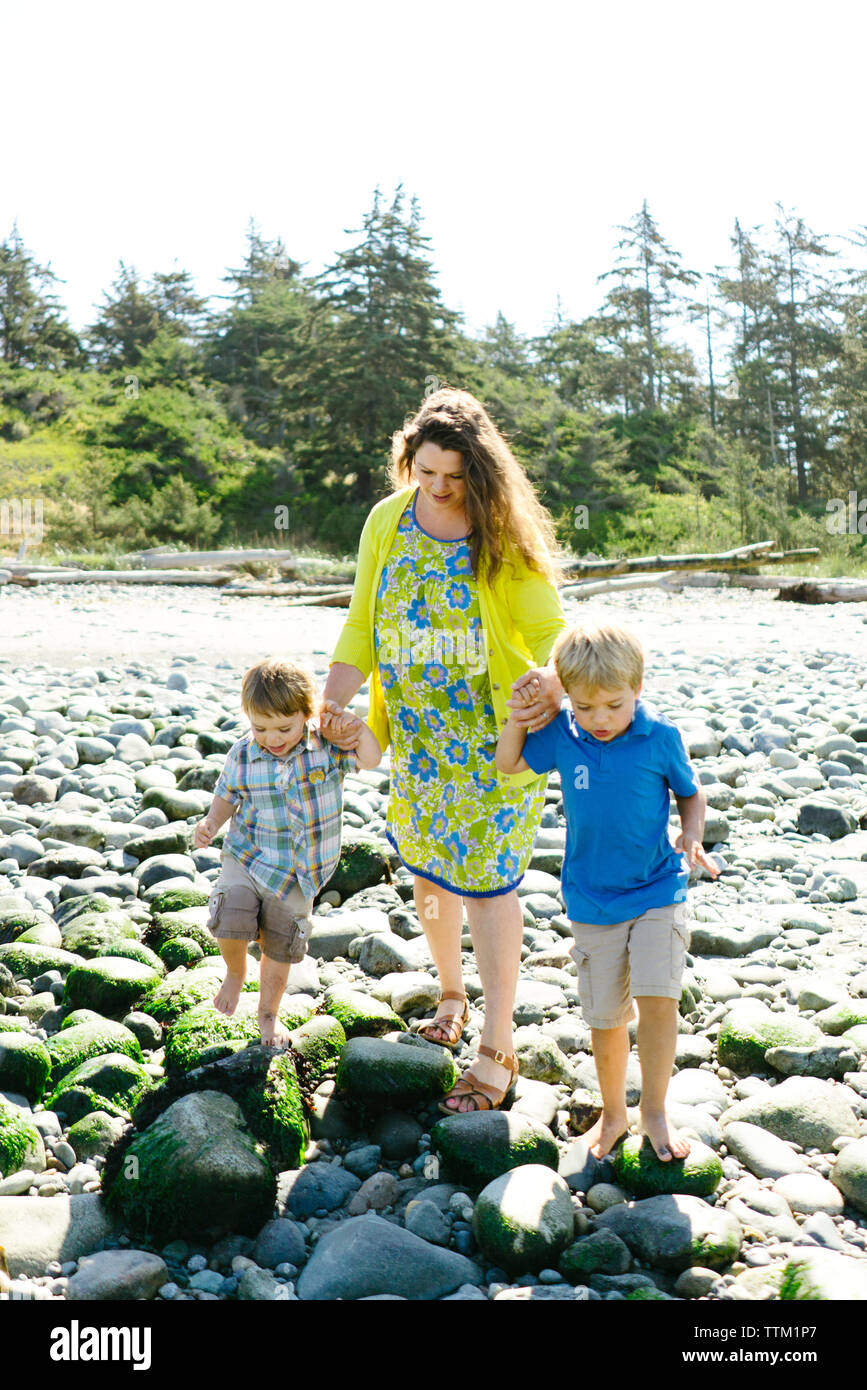 Mother holding son's hands while walking on rocks at beach against clear sky during sunny day Stock Photo