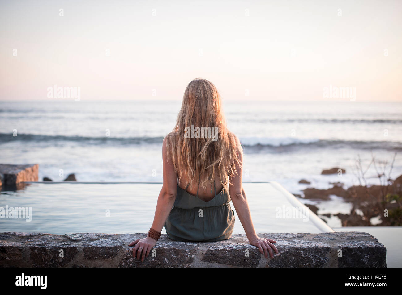 Rear view of woman sitting on retaining wall at artificial pond against sea and clear sky Stock Photo